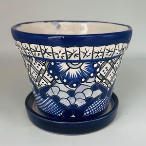 May include: A blue and white ceramic flower pot with a matching saucer. The pot features a detailed floral and geometric pattern in shades of blue, white, and gray. The rim and saucer are a solid, glossy blue.