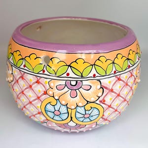 May include: A round, hand-painted ceramic planter with a vibrant floral design. The planter features a lavender rim, orange and yellow floral patterns, and a pink diamond lattice pattern. The interior is a neutral beige color.