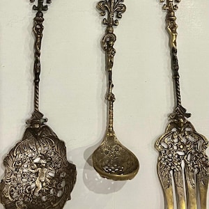 May include: A set of three brass utensils including a large perforated spoon, a smaller ladle, and a three-pronged fork. Each piece features an ornate handle with a figure and a fleur-de-lis top, with intricate cut-out designs on the bowls.