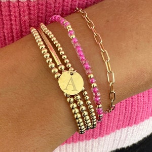 May include: A gold chain bracelet with a charm that says "A" and a pink beaded bracelet. The bracelets are worn on a person's wrist.