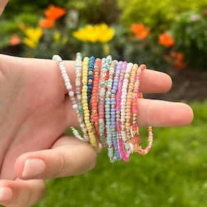 May include: A collection of colorful beaded bracelets in various shades, including white, yellow, orange, pink, blue, and gold. The bracelets are held in a hand against a blurred background of green foliage and flowers. Each bracelet appears to be approximately 7 inches in circumference.