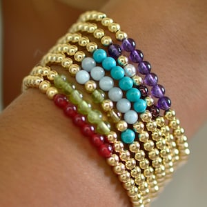 May include: A collection of beaded bracelets in various colors, including gold, red, green, light blue, turquoise, and purple. The bracelets are stacked on a wrist, showcasing the different gemstone beads.