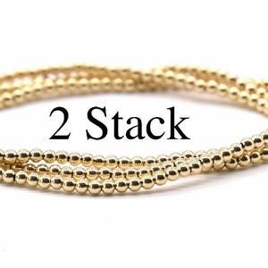 May include: Two gold beaded bracelets stacked on top of each other. The bracelets are made of small, round beads and are a simple, elegant design.