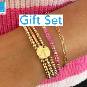 May include: A gold gift set of bracelets with a charm that says "A". The bracelets are layered on a person's wrist. The bracelets are a beaded bracelet, a chain bracelet, and a beaded bracelet with pink beads.