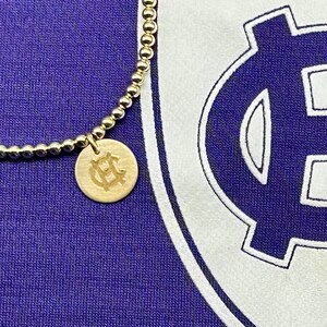 May include: Two gold necklaces with a charm featuring a monogrammed 'HC' on a purple background. The charms are hanging from a chain and a beaded bracelet. A white shield with a blue 'HC' monogram is in the background.