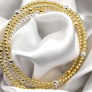 May include: A set of three gold and silver beaded bracelets. The bracelets are layered on top of each other and are made of small, round beads.