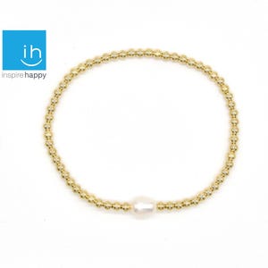 May include: Gold beaded bracelet with a single white pearl. The bracelet is made of small, round, gold-colored beads and a central, oval-shaped pearl. The bracelet is displayed on a white background. The logo "ih inspire happy" is in the upper left corner.