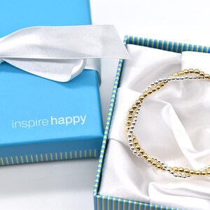 May include: A gold and silver beaded bracelet rests inside a blue gift box with a white satin lining. The box has a white ribbon tied in a bow. The text "inspire happy" is printed on the box.