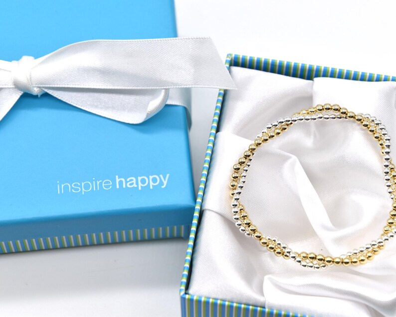 May include: A gold and silver beaded bracelet rests inside a blue gift box with a white satin lining. The box has a white ribbon tied in a bow. The text "inspire happy" is printed on the box.