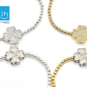 May include: Four beaded bracelets with clover charms. Two bracelets are silver-toned, and two are gold-toned. Each clover charm is encrusted with small, clear stones. The bracelets are displayed against a white background. The logo "ih inspire happy" is in the upper left corner.