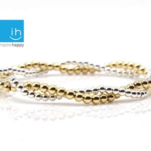 May include: A bracelet with a twisted design, featuring alternating rows of gold and silver-colored beads. The bracelet is displayed on a white surface. The logo "inspire happy" is in the upper left corner.