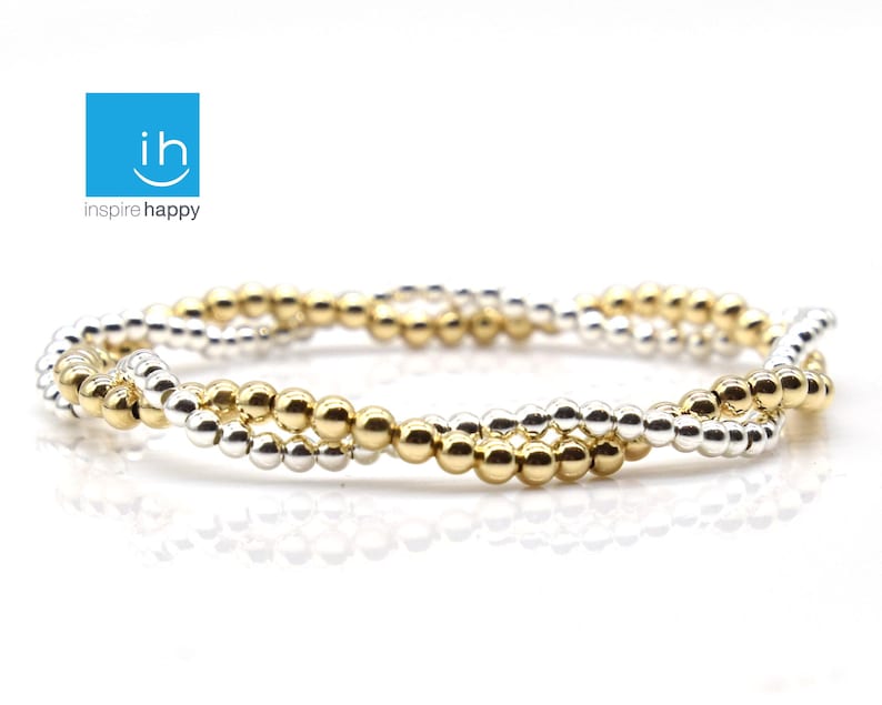 May include: A bracelet with a twisted design, featuring alternating rows of gold and silver-colored beads. The bracelet is displayed on a white surface. The logo "inspire happy" is in the upper left corner.