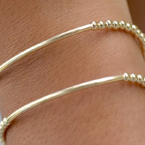 May include: Two gold bangle bracelets with a beaded design. The bracelets are stacked on a person's wrist.