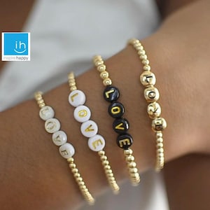 May include: Four gold beaded bracelets spelling out the word "LOVE". The bracelets feature white and black letter beads. The bracelets are on a person's arm.