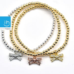 May include: Three beaded bracelets with a bow charm. The bracelets are silver, gold, and rose gold. Each bow charm has clear rhinestones.