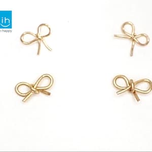 May include: Four small, gold-colored bow-shaped charms are arranged on a white background. The charms are made of thin wire, with a simple, elegant design. The upper left corner features a blue square with the text "ih inspire happy".