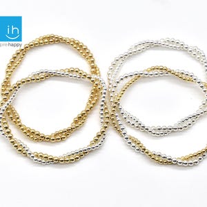 May include: A collection of beaded bracelets in gold and silver tones. The bracelets are made of small, round beads strung together to form multiple strands. The bracelets are displayed on a white surface. The logo "ih inspire happy" is in the top left corner.