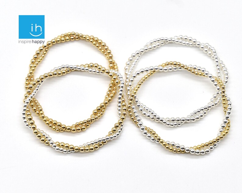 May include: A collection of beaded bracelets in gold and silver tones. The bracelets are made of small, round beads strung together to form multiple strands. The bracelets are displayed on a white surface. The logo "ih inspire happy" is in the top left corner.