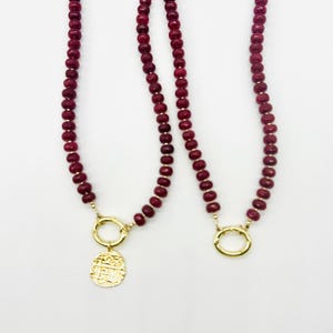 May include: Two necklaces featuring strands of small, round, deep red beads. Each necklace includes a gold-toned circular clasp. One necklace features a round, textured gold-toned pendant with an embossed design, while the other has only the clasp.