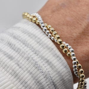 May include: Two beaded bracelets, one gold and one silver, are worn on a wrist. The bracelets are made of small, round beads. The wrist is covered by a white and gray striped sleeve.
