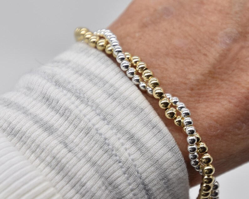 May include: Two beaded bracelets, one gold and one silver, are worn on a wrist. The bracelets are made of small, round beads. The wrist is covered by a white and gray striped sleeve.