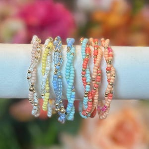 May include: A collection of beaded bracelets in various colors, including pink, yellow, blue, and coral. Each bracelet features a twisted design with small, gold-colored beads interspersed throughout. The bracelets are displayed on a white surface against a blurred floral background.