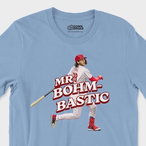 May include: A light blue short-sleeve t-shirt featuring a graphic of a baseball player in a red and white pinstripe uniform swinging a bat. The text "MR BOHM-BASTIC" is printed in bold red letters across the center of the graphic design.