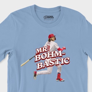 May include: A light blue t-shirt featuring a graphic of a baseball player in a white pinstripe uniform with a red helmet and bat. The text MR BOHM-BASTIC is printed in large red letters across the center. The shirt has a crew neck and short sleeves.