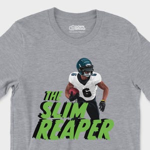 Puede incluir: Una camiseta gris jaspeado de manga corta con el gráfico de un jugador de fútbol americano con camiseta blanca número 6, sosteniendo un balón. Debajo del jugador, el texto THE SLIM REAPER está impreso en una fuente verde lima desgastada.