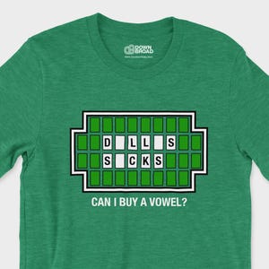 May include: A heather green short-sleeved t-shirt featuring a graphic of a game board with the letters D, I, L, L, S, S, C, K, S in white tiles. Below the board, the text CAN I BUY A VOWEL? is printed in white capital letters.