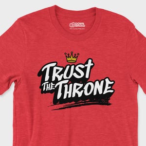 May include: A heather red short-sleeve t-shirt featuring the text "TRUST THE THRONE" in bold white letters with black shadowing. Above the text is a small yellow crown graphic. The shirt has a crew neckline and a brand logo printed inside the collar.