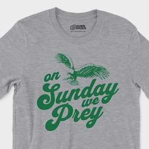 以下が含まれることがあります： グレーのヘザーカラーの半袖Tシャツ。飛行するワシの緑色のグラフィックと、筆記体で「on Sunday we Prey」という文字がプリントされています。クルーネックで、襟の内側には小さなロゴが印刷されています。