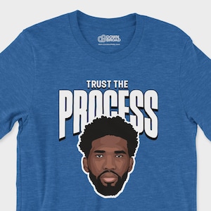 May include: A heathered royal blue short-sleeve t-shirt featuring a graphic illustration of a bearded person with dark hair. Above the illustration, the text reads "TRUST THE PROCESS" in bold white capital letters with a black outline.