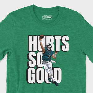 Op de afbeelding: Een heidegroen t-shirt met korte mouwen en een grafische illustratie van een footballspeler in een donkergroen shirt met het nummer 1, die een football gooit. De tekst "HURTS SO GOOD" staat in grote witte letters achter de speler.