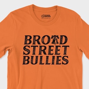 K&ouml;nnte beinhalten: Ein orangefarbenes T-Shirt mit kurzen &Auml;rmeln und dem schwarzen Schriftzug "BROAD STREET BULLIES", wobei ein Liberty Bell-Symbol den Buchstaben "A" in "BROAD" ersetzt. Das Shirt hat einen Rundhalsausschnitt, ein kleines Logo am linken &Auml;rmel und ein Innenetikett.