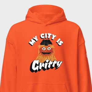 Può includere: Una felpa con cappuccio color arancione brillante con la grafica di una creatura arancione pelosa che indossa un casco da hockey nero. Il testo sopra la creatura recita "MY CITY IS" e sotto recita "Gritty" in lettere bianche in grassetto.