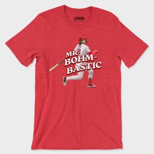 May include: A heather red short-sleeve t-shirt featuring a graphic of a baseball player in a white pinstripe uniform swinging a bat. The text "MR BOHM-BASTIC" is printed in bold white letters with a dark red outline across the center of the chest.