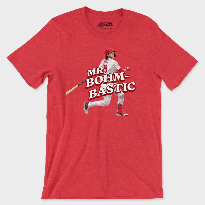 May include: A heather red short-sleeve t-shirt featuring a graphic of a baseball player in a white pinstripe uniform swinging a bat. The text "MR BOHM-BASTIC" is printed in bold white letters with a dark red outline across the center of the chest.