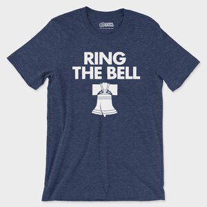 K&ouml;nnte beinhalten: Ein kurz&auml;rmeliges T-Shirt in meliertem Marineblau mit dem wei&szlig;en Schriftzug "RING THE BELL" &uuml;ber einer Grafik der Liberty Bell. Das T-Shirt hat einen Rundhalsausschnitt und ein Markenlogo im inneren Kragenbereich.