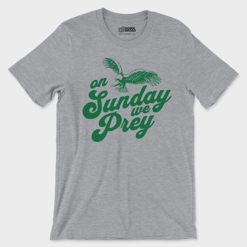 以下が含まれることがあります： グレーのヘザーカラーの半袖Tシャツ。飛行するワシの緑色のグラフィックと、筆記体で「on Sunday we Prey」という文字がプリントされています。クルーネックで、襟の内側には小さなロゴが印刷されています。