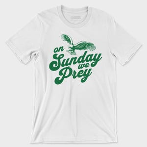 以下が含まれることがあります： 白い半袖Tシャツで、胸の中央に飛んでいるワシの緑色のグラフィックと、太字の筆記体で「on Sunday we Prey」という文字がプリントされています。クルーネックで、標準的なフィット感のTシャツです。