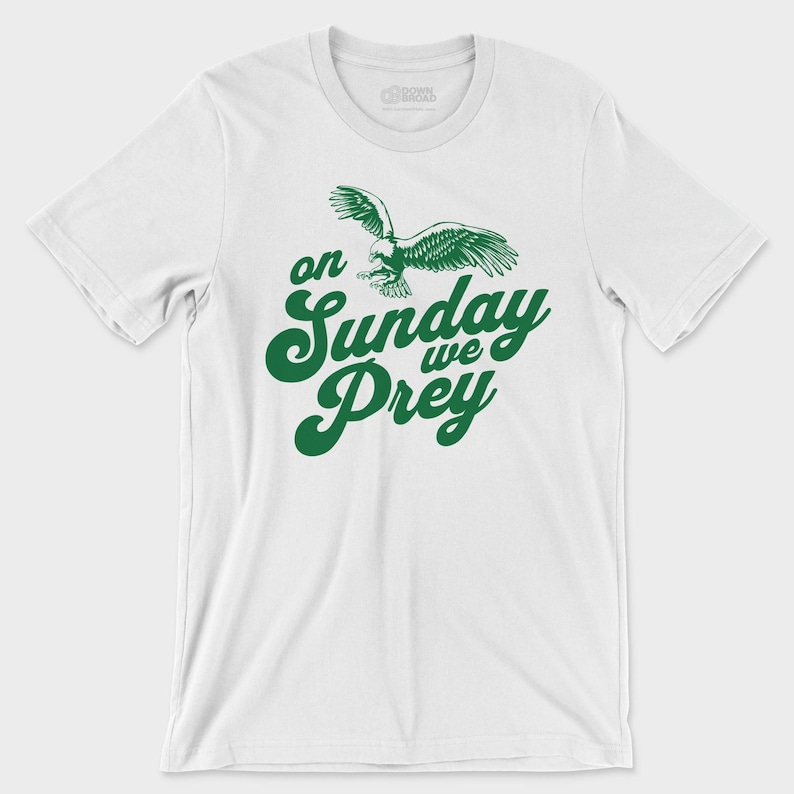 以下が含まれることがあります： 白い半袖Tシャツで、胸の中央に飛んでいるワシの緑色のグラフィックと、太字の筆記体で「on Sunday we Prey」という文字がプリントされています。クルーネックで、標準的なフィット感のTシャツです。