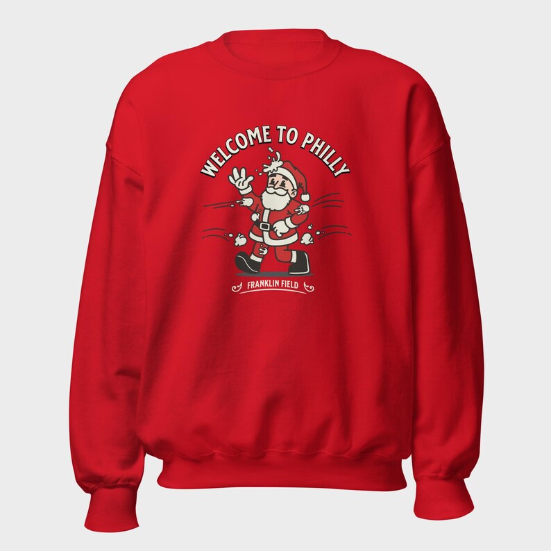 May include: A bright red long-sleeve sweatshirt featuring a graphic of Santa Claus being hit by snowballs. The text above reads "WELCOME TO PHILLY" and the text below reads "FRANKLIN FIELD". The design includes white, black, and grey accents.