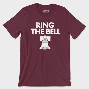 K&ouml;nnte beinhalten: Ein kastanienbraunes T-Shirt mit kurzen &Auml;rmeln und Rundhalsausschnitt, das den wei&szlig;en Schriftzug "RING THE BELL" &uuml;ber einer grafischen Abbildung der Liberty Bell zeigt. Das Innenetikett am Kragen zeigt das "DOWN BROAD"-Logo.