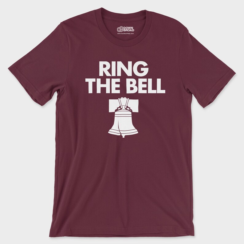 K&ouml;nnte beinhalten: Ein kastanienbraunes T-Shirt mit kurzen &Auml;rmeln und Rundhalsausschnitt, das den wei&szlig;en Schriftzug "RING THE BELL" &uuml;ber einer grafischen Abbildung der Liberty Bell zeigt. Das Innenetikett am Kragen zeigt das "DOWN BROAD"-Logo.