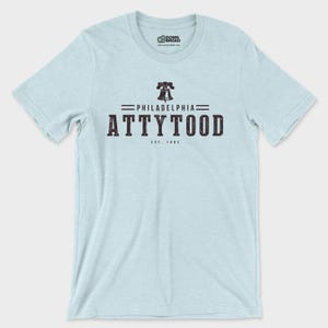 Puede incluir: Una camiseta de manga corta de color azul claro con un gr&aacute;fico oscuro en el pecho. El dise&ntilde;o incluye un icono de la Liberty Bell sobre el texto PHILADELPHIA ATTYTOOD y EST. 1682. El logotipo de la marca DOWN BROAD aparece en el cuello.
