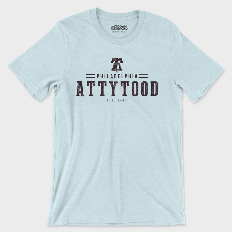 Puede incluir: Una camiseta de manga corta de color azul claro con un gr&aacute;fico oscuro en el pecho. El dise&ntilde;o incluye un icono de la Liberty Bell sobre el texto PHILADELPHIA ATTYTOOD y EST. 1682. El logotipo de la marca DOWN BROAD aparece en el cuello.