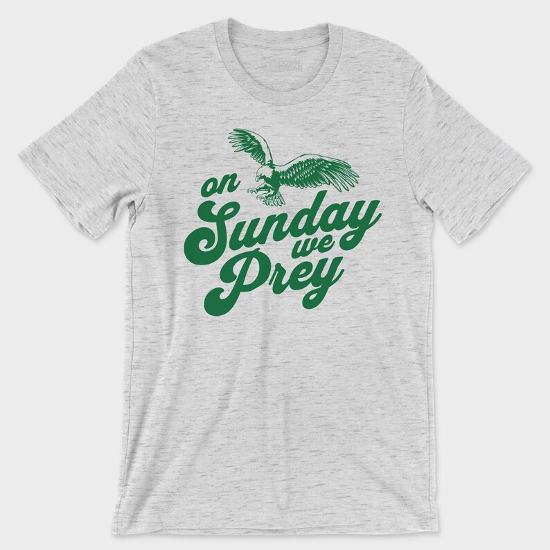 以下が含まれることがあります： ライトグレーの霜降り半袖Tシャツ。胸の中央には、飛行するワシのダークグリーンのグラフィックと、太字の筆記体で「on Sunday we Prey」というテキストがプリントされています。クルーネックのシンプルなデザインの衣類です。