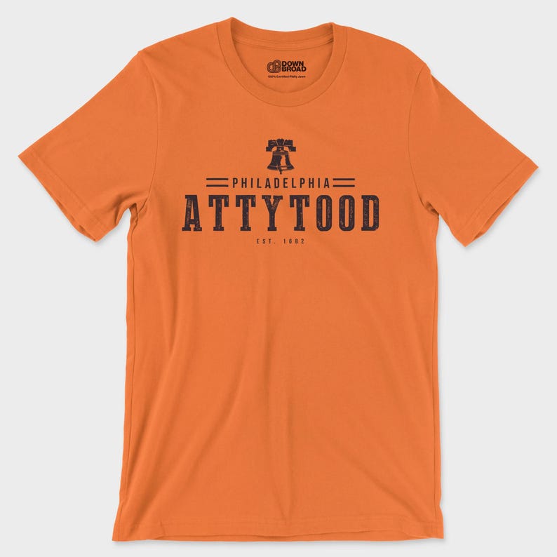 Puede incluir: Una camiseta naranja de manga corta con un gr&aacute;fico azul oscuro en el pecho. El gr&aacute;fico incluye una ilustraci&oacute;n de la Liberty Bell, el texto "PHILADELPHIA", la palabra "ATTYTOOD" en letras grandes y el texto "EST. 1882" debajo.