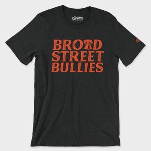 K&ouml;nnte beinhalten: Ein anthrazitfarbenes Kurzarm-T-Shirt mit dem orangefarbenen Schriftzug "BROAD STREET BULLIES", wobei ein Liberty Bell-Symbol den Buchstaben A in Broad ersetzt. Das T-Shirt hat einen Rundhalsausschnitt und ein kleines Logo am Innenkragen und am linken &Auml;rmel.